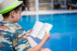 A young boy relaxes by the pool, reading a book in summer attire at a resort.