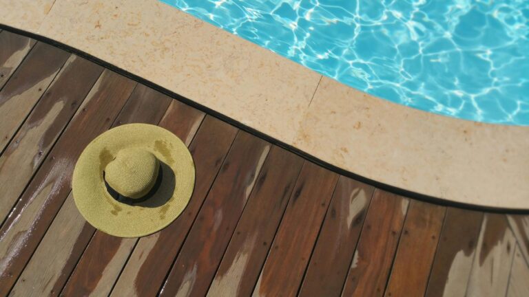 A straw hat rests on a wooden deck beside a sparkling blue swimming pool, perfec